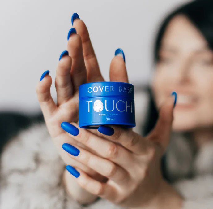 Ženské ruce držíci modrej box s nápisem značky TOUCH, který obsahuje krycí lak. Nehty jsou modré barvy.