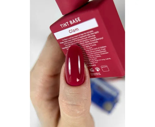 Hand with red nail polish holding a red product packaging labeled 'Tint Base Glam'.